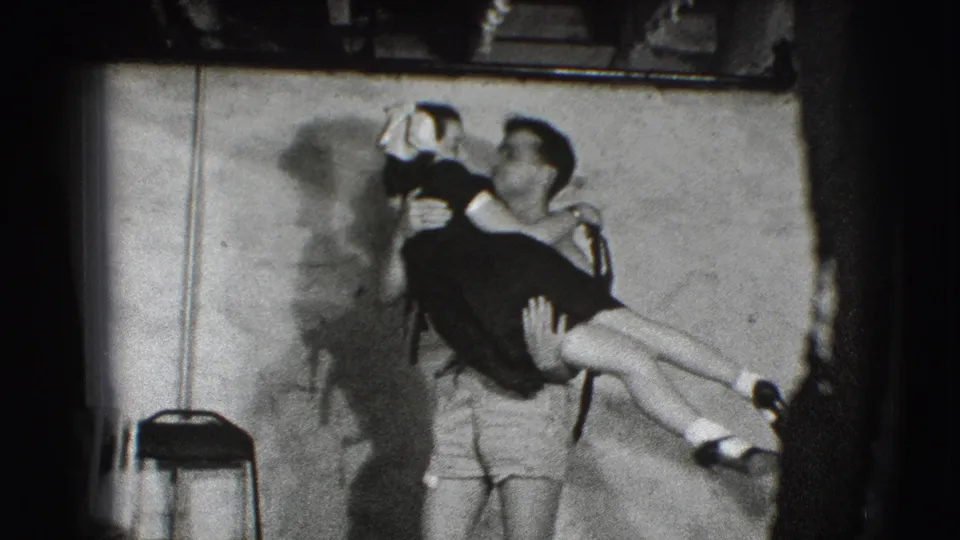 In the small 1941 Joliet room, a young man lifts a woman in a dark dress as another person stands close behind him.