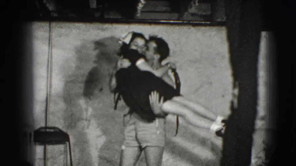 A young man carries a woman in a dark dress across the small Joliet interior, with a chair, textured wall, and heavy film-edge shadow visible.