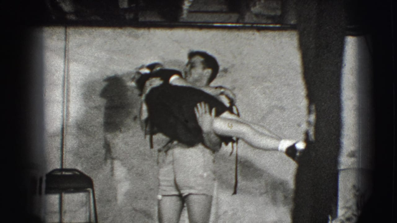 Man in shorts lifts a woman in a dark dress against a plain indoor wall.