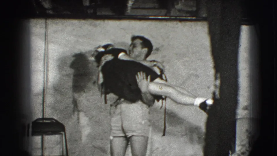 In a 1941 Joliet home-movie frame, a man in shorts holds and lifts a woman in a dark dress in a plain interior.