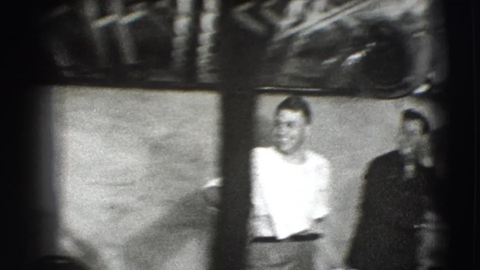 A 1941 Joliet home-movie frame shows a man in a white shirt standing near another adult in a dark shirt inside a shadowed room.