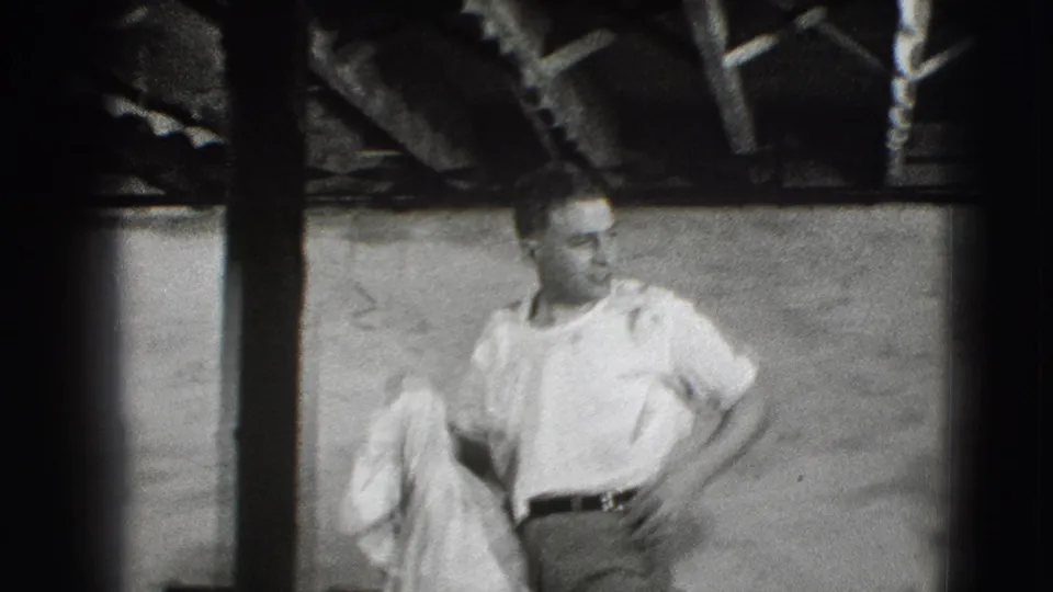 The man in a white shirt holds a light cloth near a chair, with exposed beams and soft 8mm grain visible in the 1941 Joliet footage.