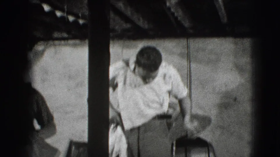 A man bends over a light cloth near a chair in a dim 1941 Joliet home interior, framed through dark vertical film or room edges.