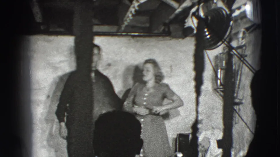 The woman faces upward beside the man in the Joliet studio interior, with a large lamp, stand, and overhead structure visible.
