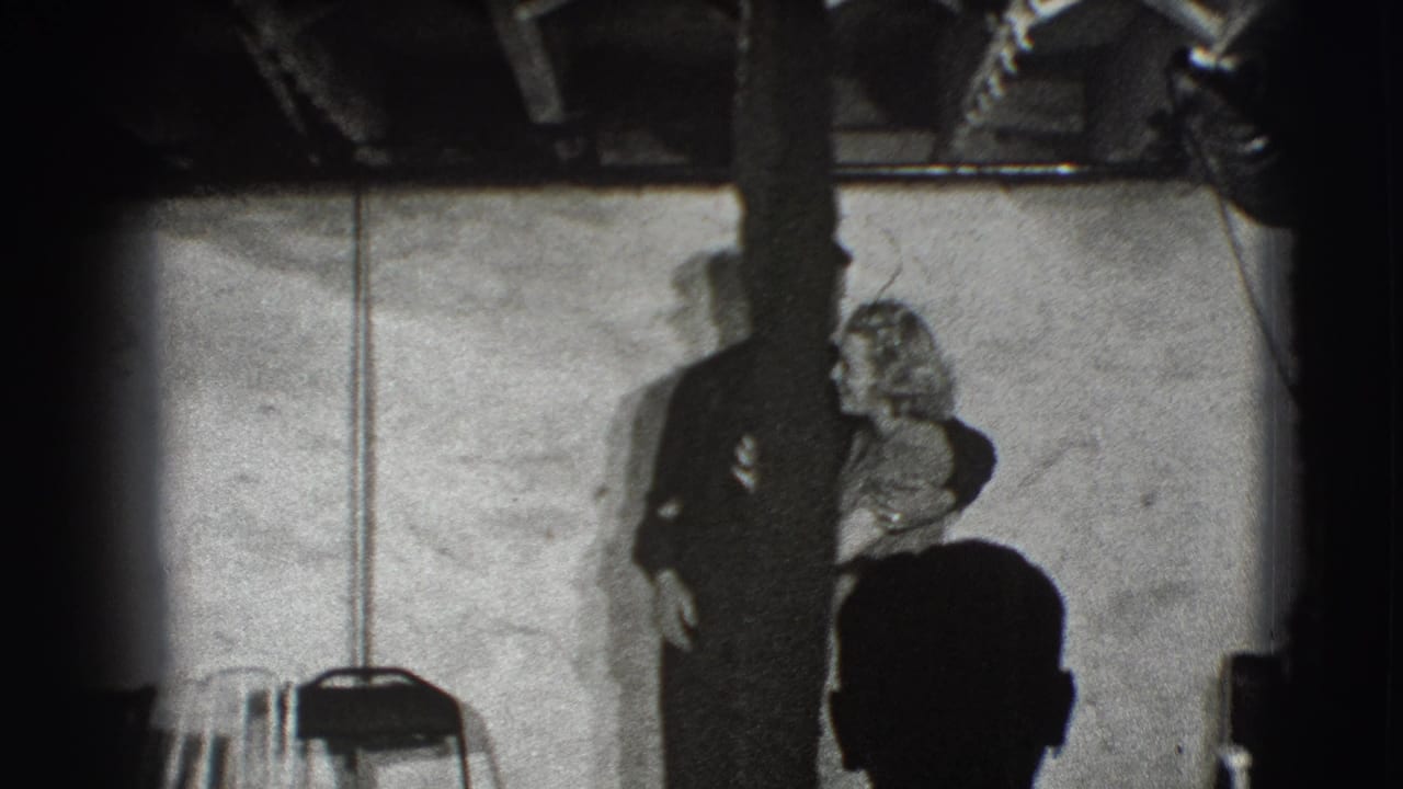 Adult man and woman stand arm-in-arm indoors, partly obscured by a vertical post.