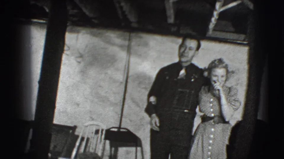 The 1941 Joliet home-movie frame shows a smiling adult couple walking arm-in-arm toward the camera indoors.