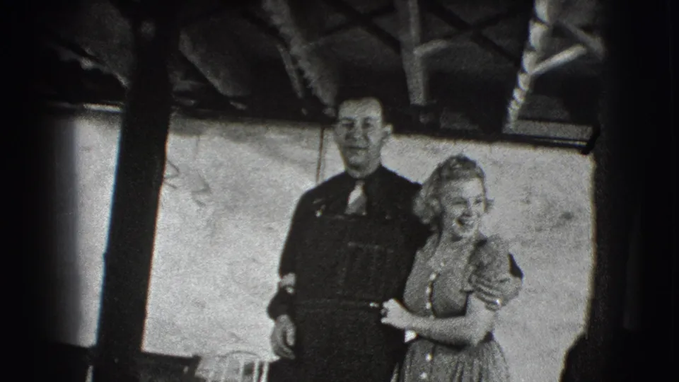 In the Joliet 1941 frame, the adult couple smiles together indoors, with wall panels and overhead structure behind them.