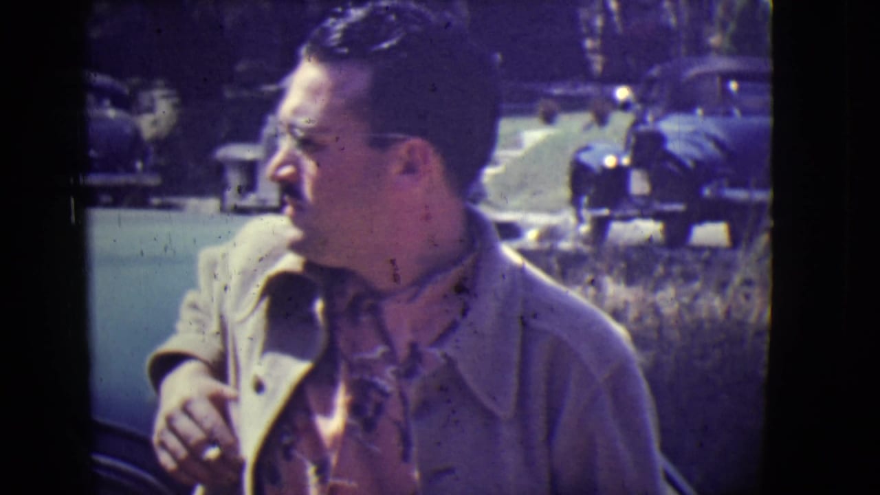 Man in a light jacket looks to the side outdoors with parked vintage cars behind him.