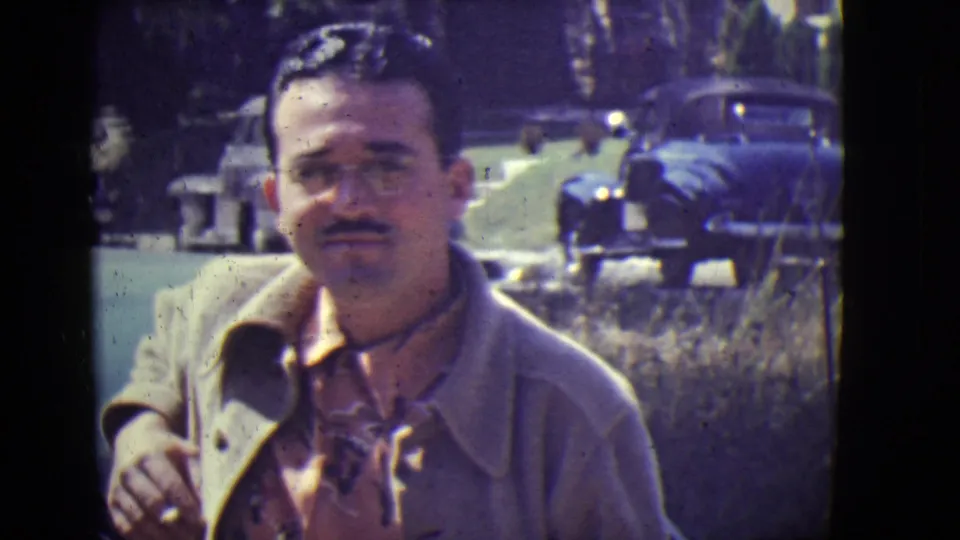 Adult man faces the camera in 1946 East Orange, with parked cars and roadside grass in the background.