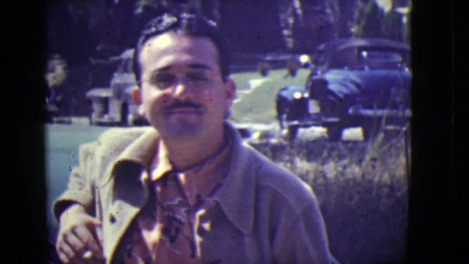 Adult man smiles toward the camera in a 1946 East Orange home-movie frame with classic cars behind him.