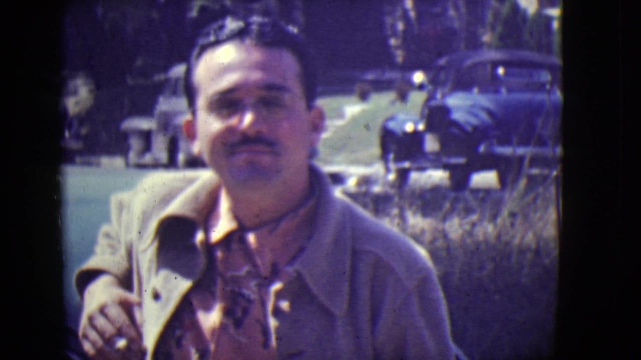 Man with moustache and glasses in a light coat stands near a car on a street.