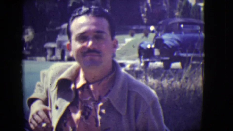 In 1946 East Orange home-movie footage, a man with moustache and glasses faces the camera near parked cars on a suburban street.