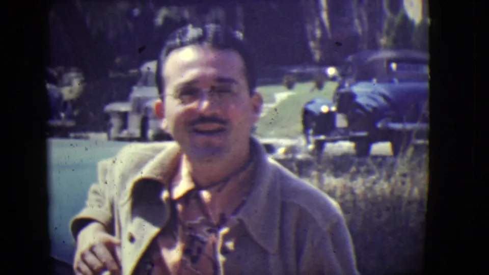The man smiles at the camera in the East Orange street scene, with mid-century parked cars and greenery visible behind him.