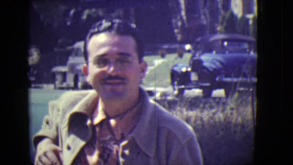 The man remains framed close to camera in 1946 East Orange footage, wearing glasses and a light coat as parked cars line the background.