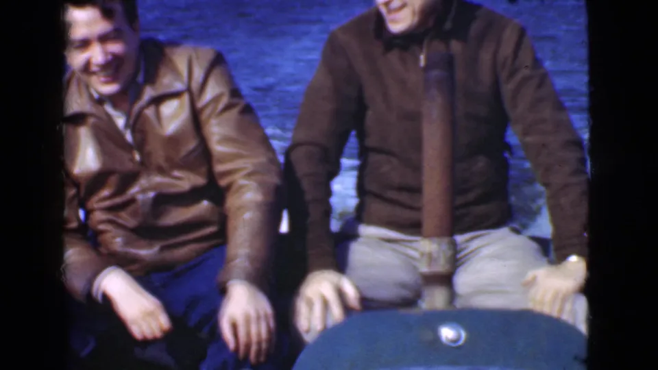 Two adult men in jackets sit on a small motorboat in Quebec home-movie footage from 1946, with open water visible behind them.