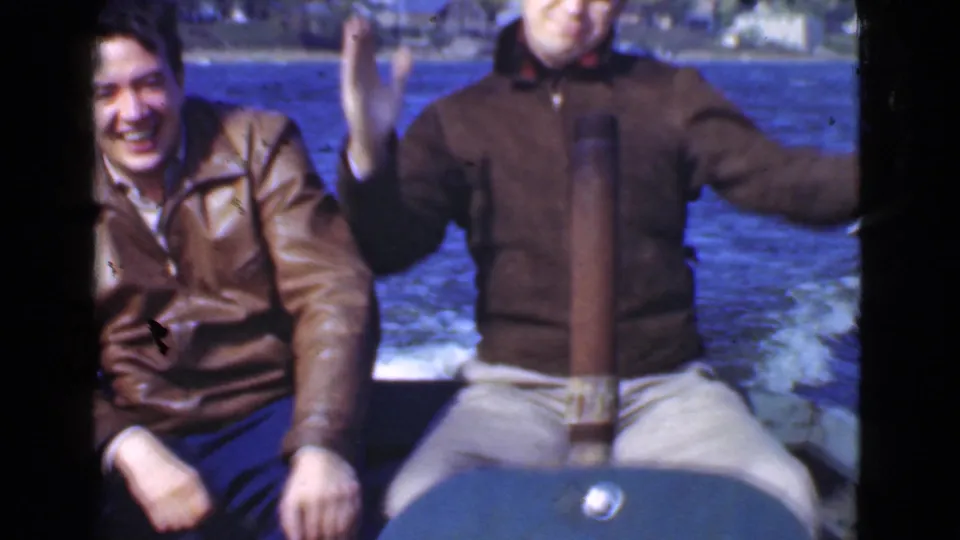 On a small motorboat in 1946 Quebec, one man laughs toward the camera while the other raises his arms with water behind them.