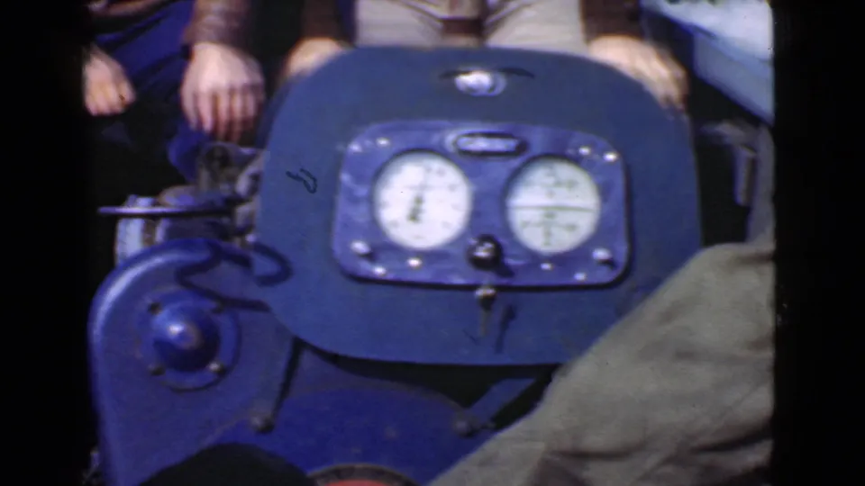 A blue motor or control housing with round gauges dominates the frame, with the seated boat riders partly visible behind it.