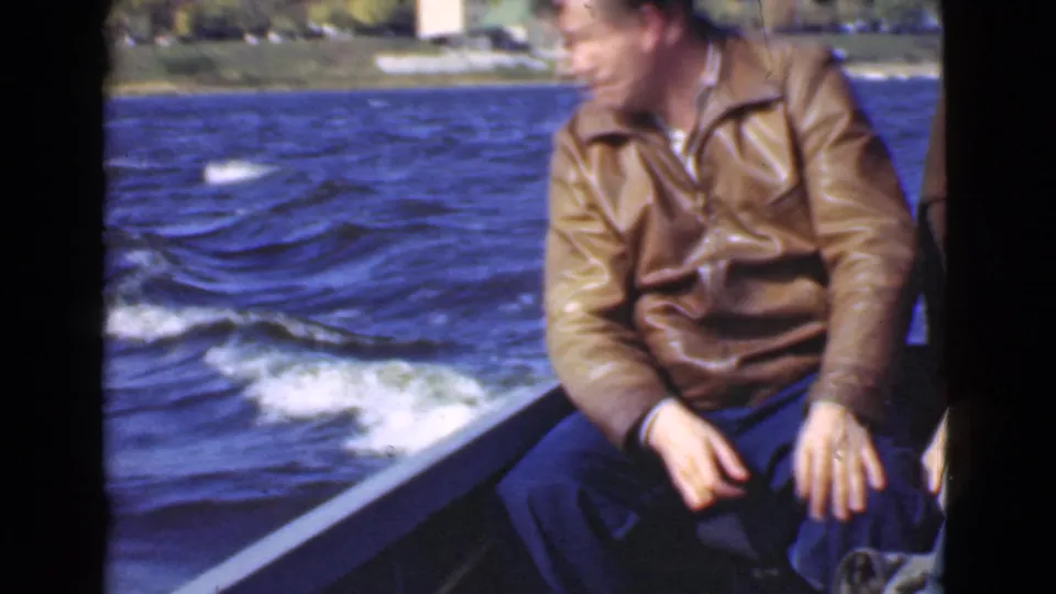 1946 Quebec 8mm frame of an adult man in a brown leather jacket seated on a motorboat as water and shoreline pass behind him.