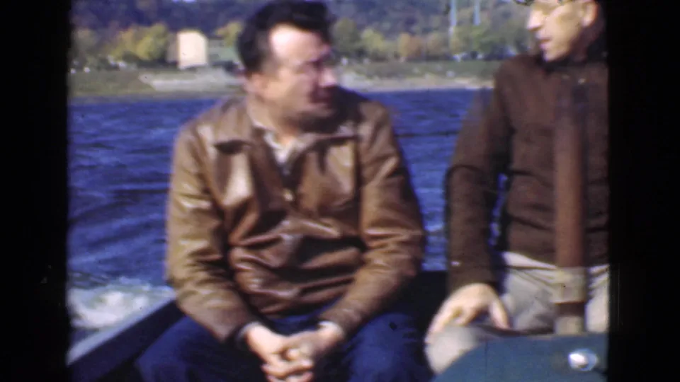 1946 Quebec 8mm frame showing two adult men seated on a moving motorboat, apparently conversing near open water and shoreline.
