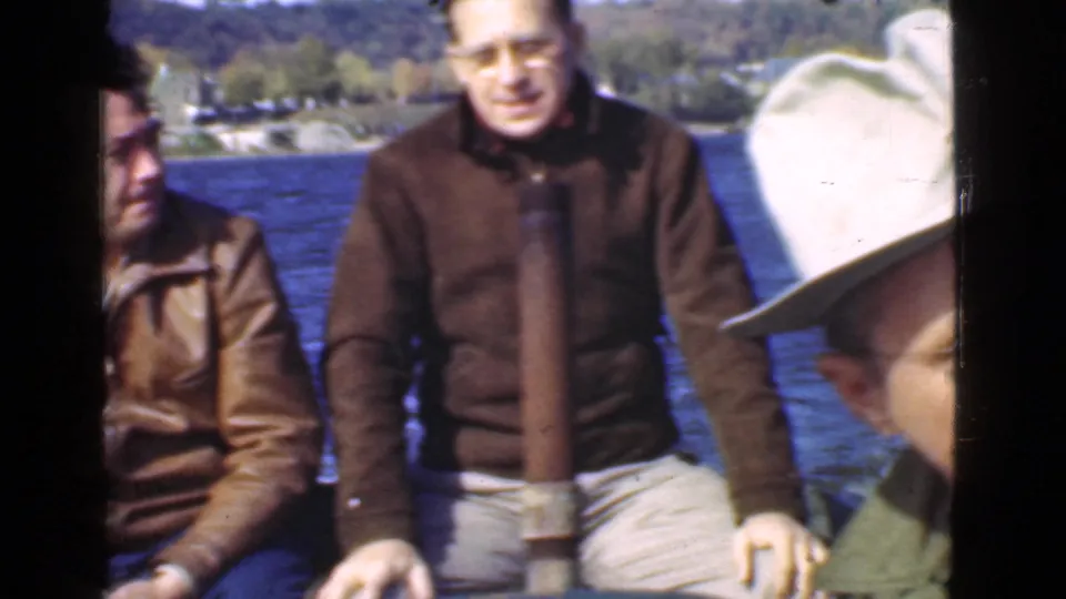 Two adult men in vintage jackets sit aboard a small boat on a Quebec lake, with wooded shoreline visible in the background.