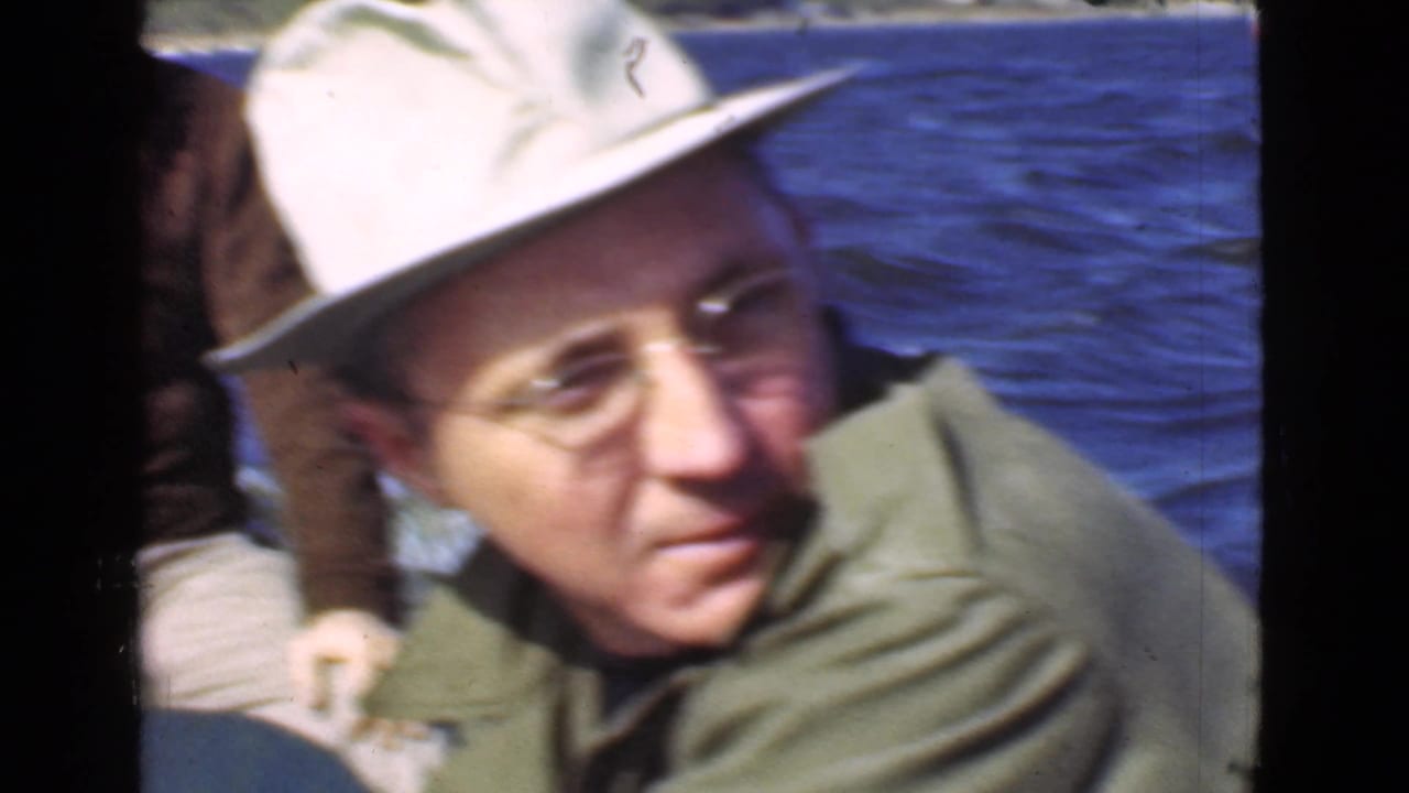 Man in a light hat and glasses sits close to the camera on a boat over blue water.