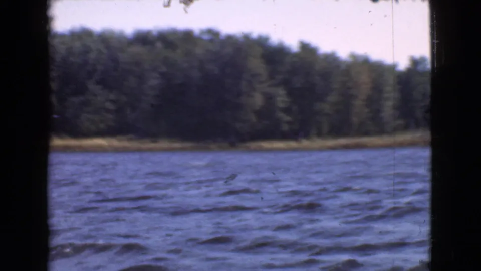 1946 Quebec 8mm frame with rippled water in front and a wooded shore under bright sky.