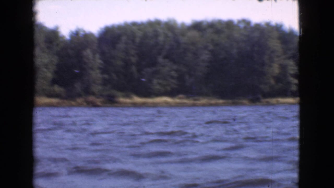 1946 Quebec 8mm view across rippling blue lake water toward a dense wooded shoreline, framed by dark gate edges.