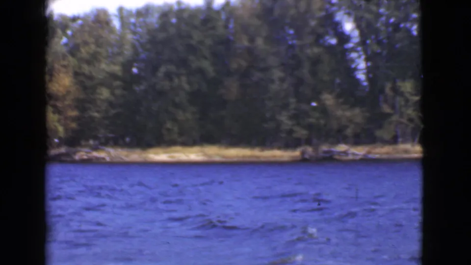 1946 Quebec 8mm view of choppy blue water with a tree-covered shoreline and narrow sandy edge in the background.