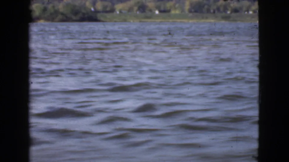 1946 Quebec home-movie frame showing lake water through dark boat structure, with a distant wooded shoreline near the top edge.