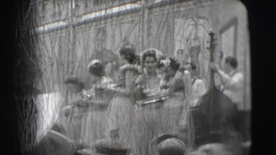 1948 Sarasota home-movie frame showing women in hula-style costumes and leis performing on stage, with audience heads in the foreground.