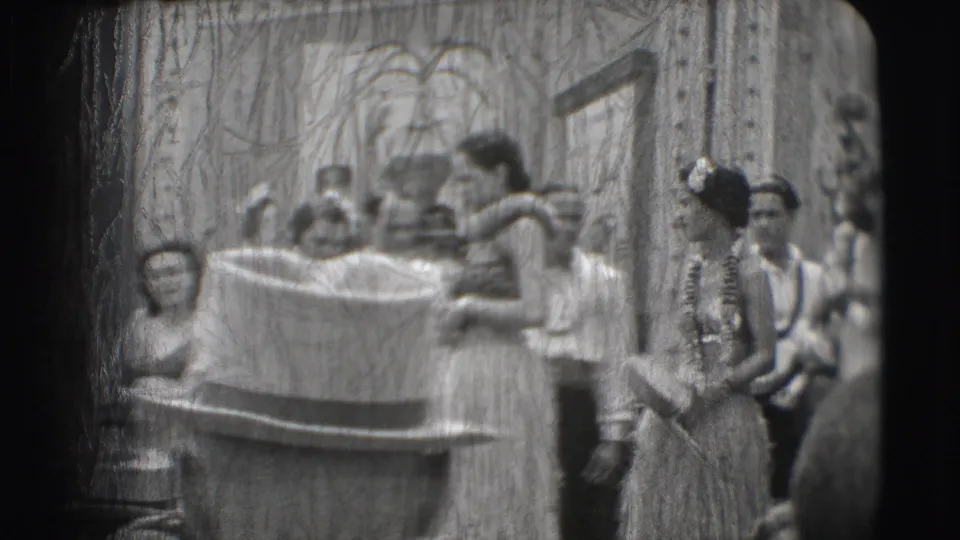 A cello player and hula-costumed performers appear on the Sarasota stage, with heavy scratches and soft 8mm focus across the frame.