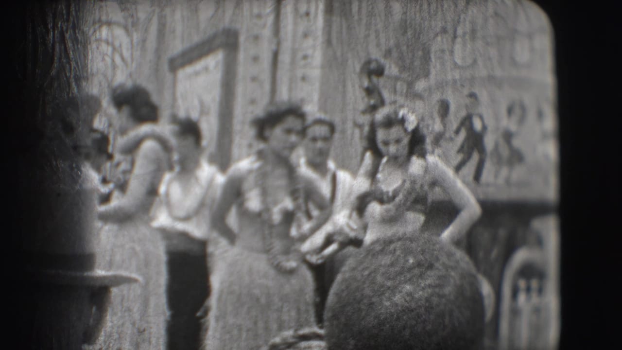 Women in grass skirts and leis stand with male performers near a mural backdrop.