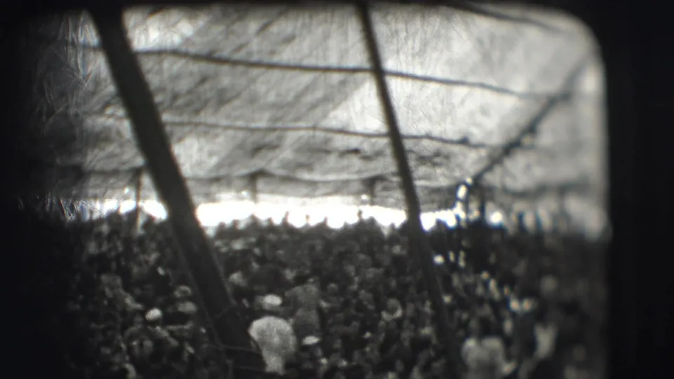 Audience view under a canvas circus tent roof, with poles crossing the frame and a bright opening beyond the crowd.