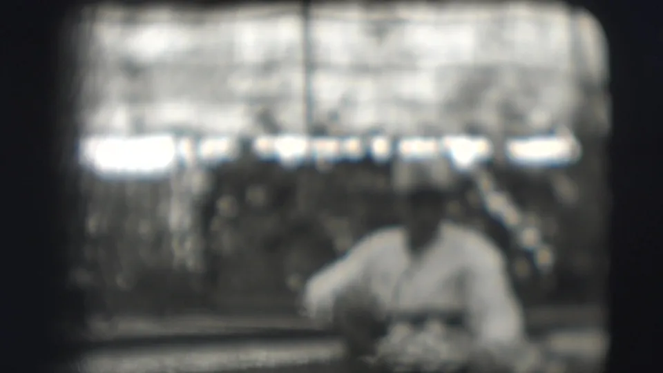 A soft 8mm frame shows the seated man at lunch, with rows of cots and the building structure visible behind him.