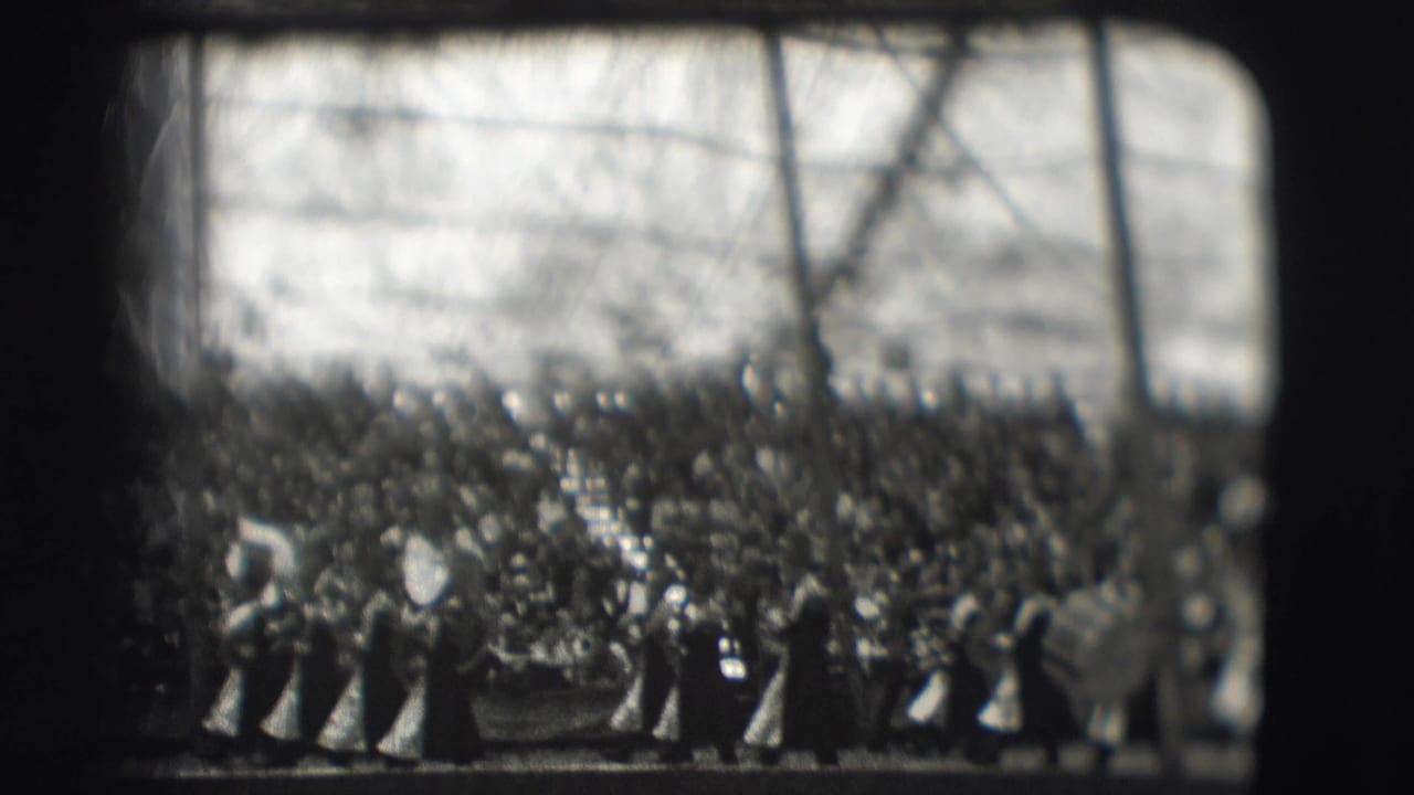 Crowd and marching band members gathered beneath tree branches in black-and-white footage.
