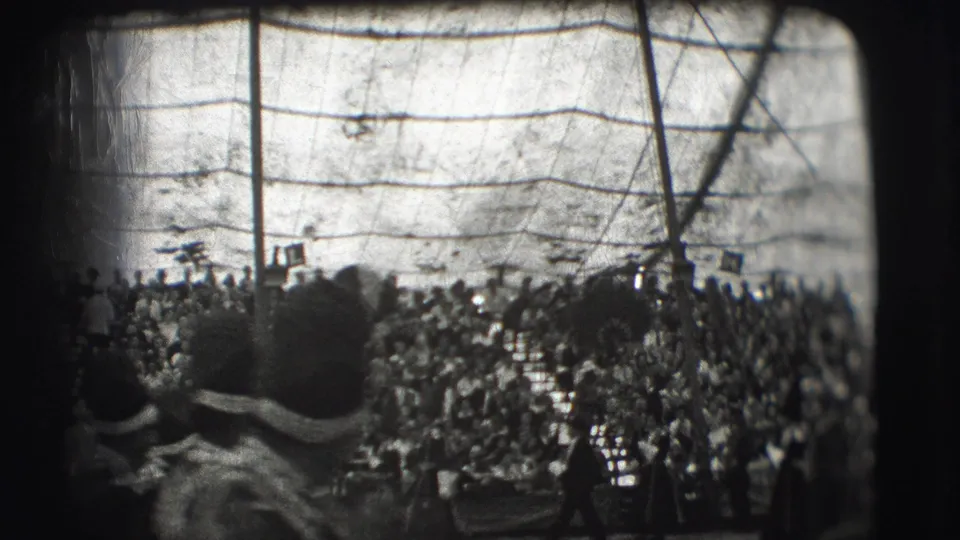 From within the crowd at the Sarasota circus, spectators in hats face a busy arena beneath visible tent or rigging lines.
