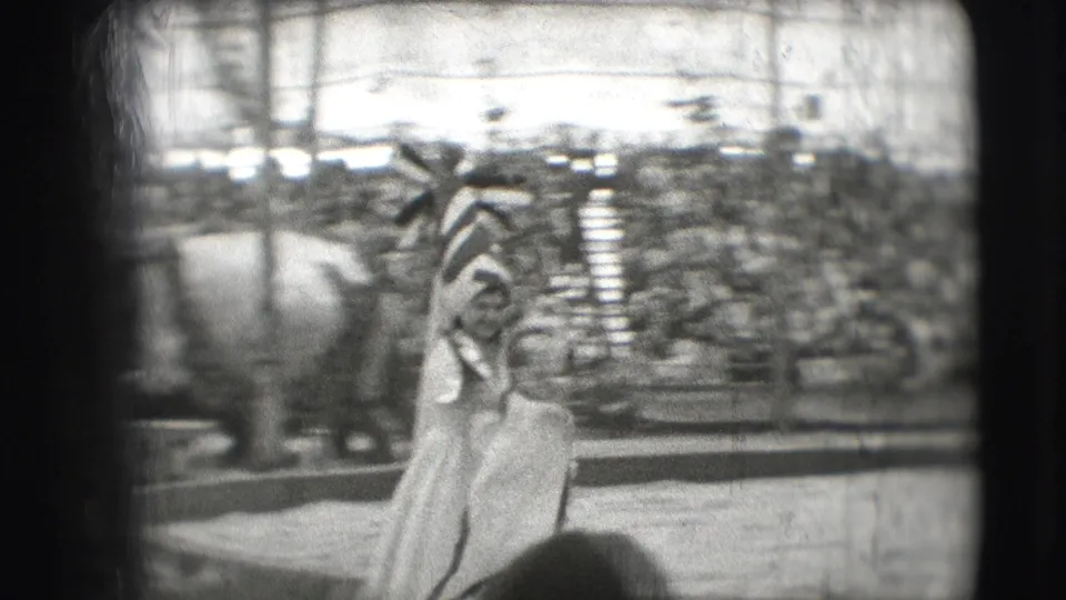 A performer in a flowing light costume stands in the ring during the 1948 Sarasota circus footage, framed by audience seating and arena barriers.