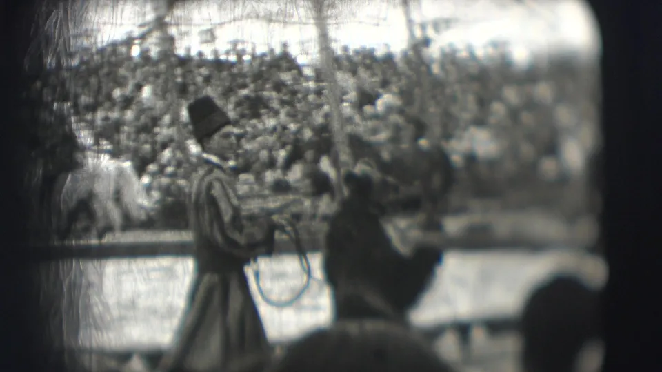 A tall-hatted performer in striped clothing faces a bear in the circus ring, with packed bleachers behind him.