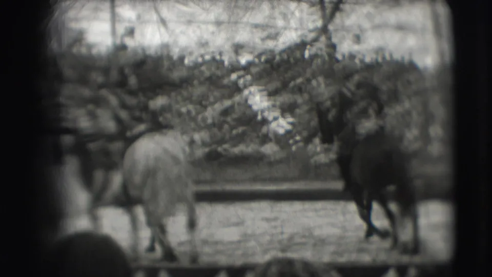 Two horses and riders pass through the circus ring while a large audience watches from the stands in the background.