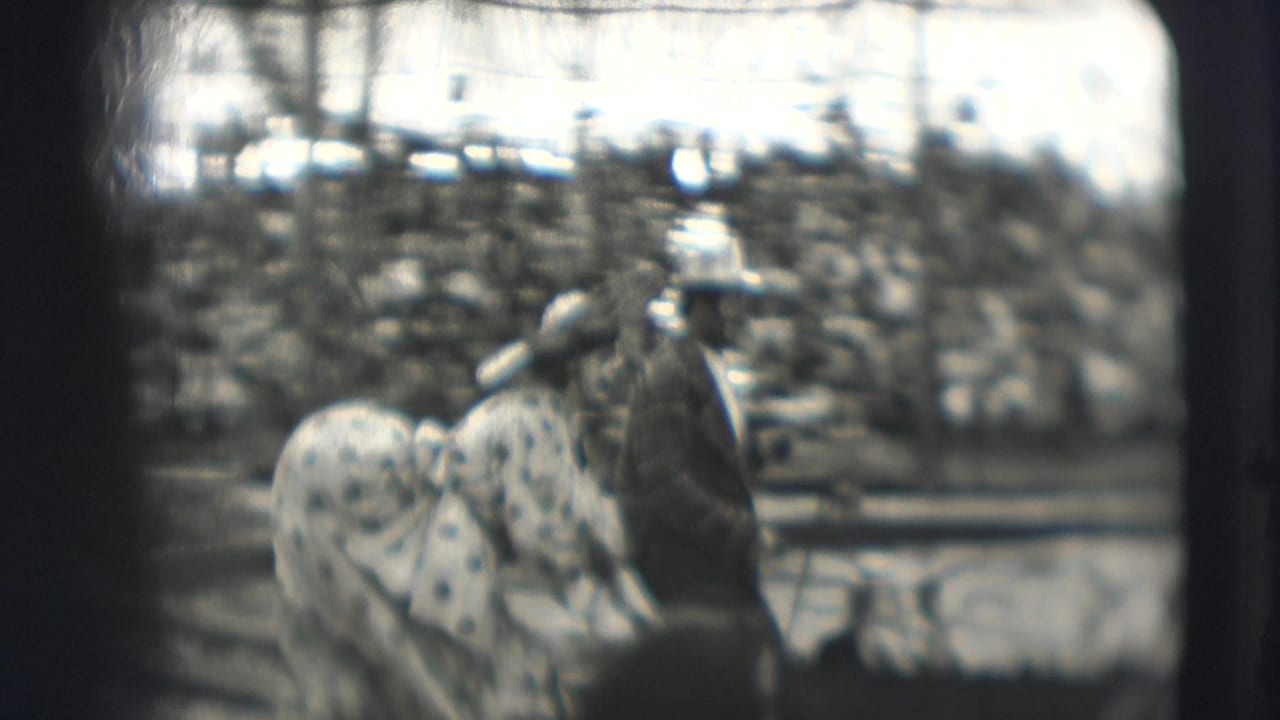 Clown pushes a baby carriage near a suited performer inside a circus tent.