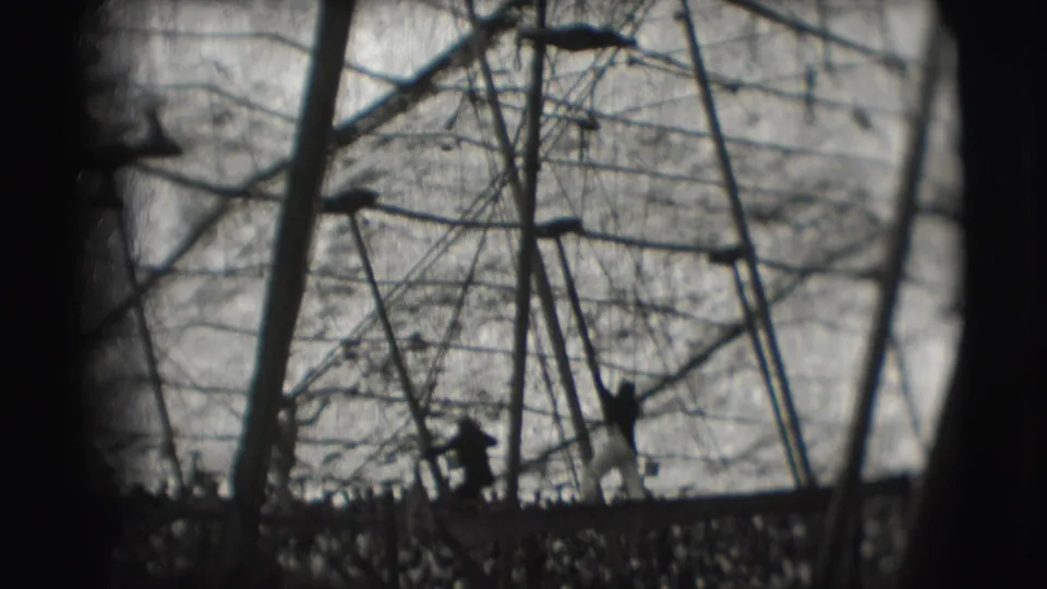 Black-and-white 8mm frame of a circus acrobat or crew figure near the lower rigging, with wires and poles crossing the sky.