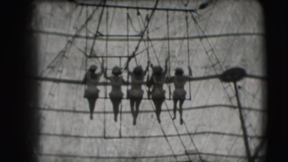 Women performers appear in silhouette on a trapeze swing at the Sarasota circus site, framed by ropes and canvas.