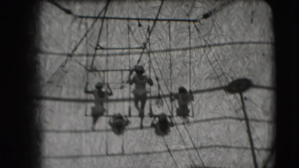 A low-angle 8mm frame shows acrobats spaced across the high bar while two performers hang below against the circus tent roof.