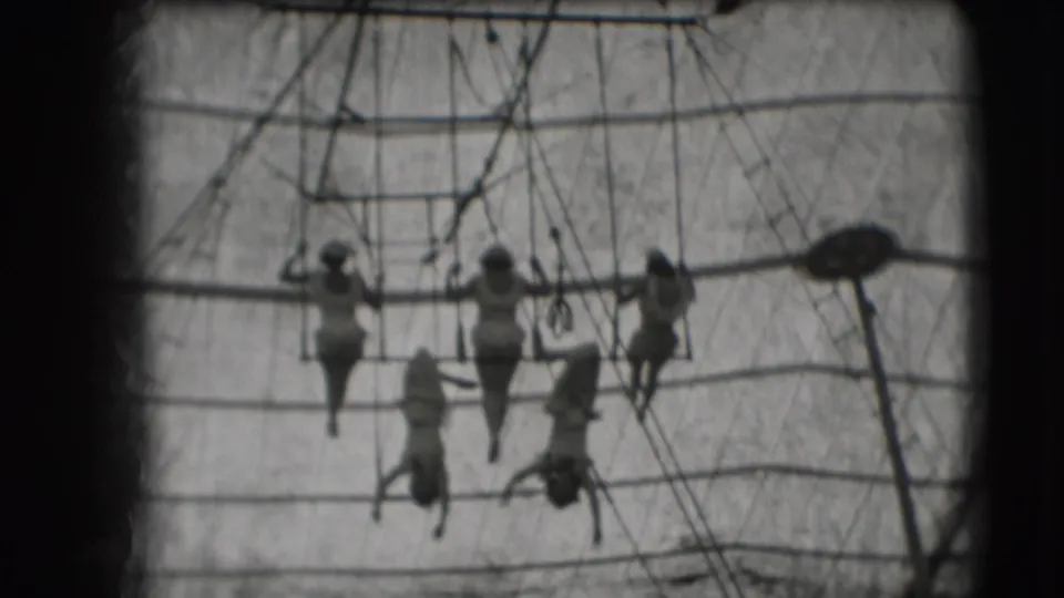 Five acrobats remain grouped around the high bar in the 1948 Sarasota circus tent, seen through grainy black-and-white 8mm film.