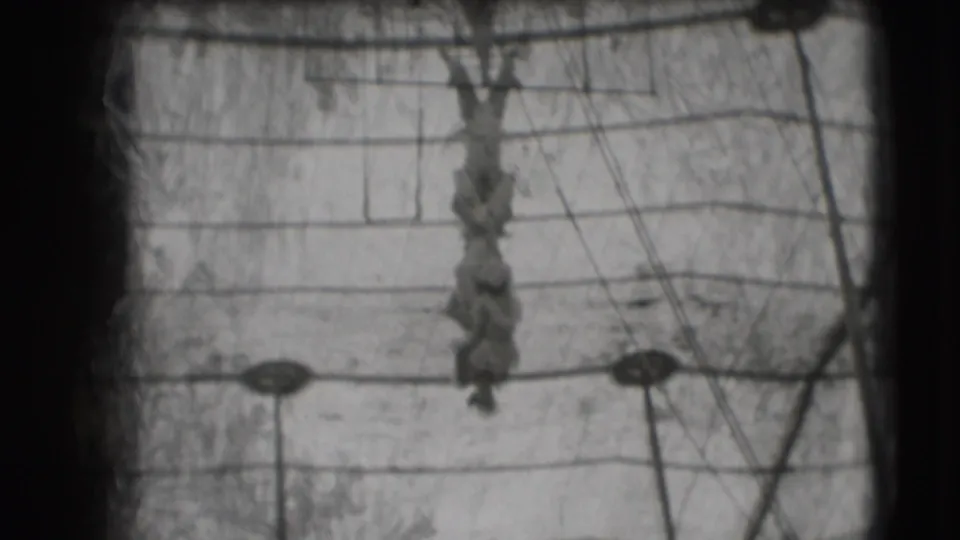 Black-and-white 8mm frame of a 1948 Sarasota trapeze act, with the performer hanging below the bar among crossing cables.