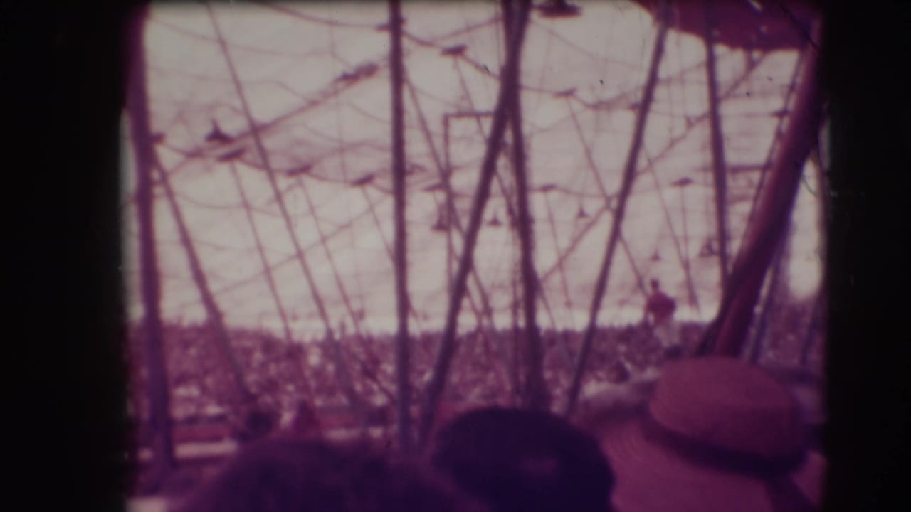 Crowded circus tent seen past poles and foreground spectators, with a distant performer on horseback.