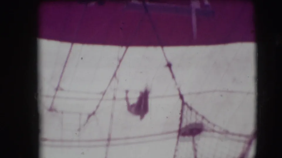 Circus tent canvas and overhead lines dominate the frame, with a small aerial performer or apparatus visible below.