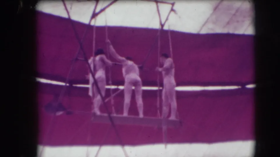 Three male acrobats in white costumes prepare on a trapeze platform inside the Sarasota circus tent, with ropes crossing the frame.