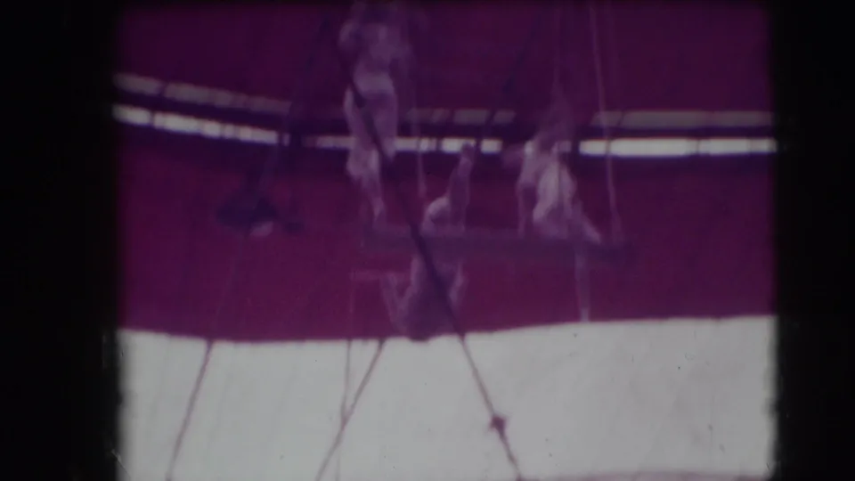 The camera drops toward the circus ring, showing rigging, a dark tent band, and blurred performers or attendants below.