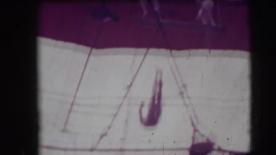 Soft, purple-tinted frame of a trapeze act with a blurred performer below tent rigging in 1948 home-movie footage.